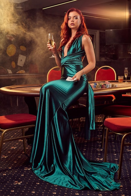 Giovane donna dai capelli rossi in un lungo abito di raso blu, con un bicchiere di champagne e carte da gioco in mano è in posa seduta su un tavolo da poker in un casinò di lusso. Passione, carte, fiches, alcol, vincita, gioco