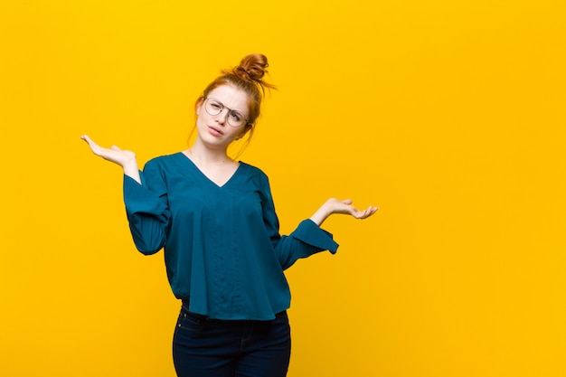 Giovane donna dalla testa rossa che sembra perplessa, confusa e stressata, chiedendosi tra diverse opzioni, sentendosi incerta contro la parete arancione