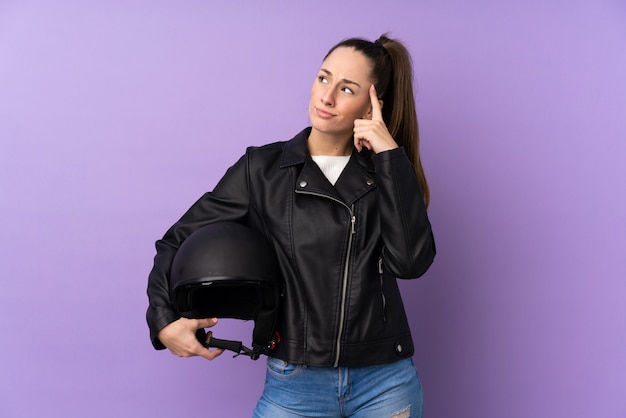 Giovane donna del brunette con un casco del motociclo sopra la parete viola isolata che ha dubbi e con l'espressione confusa del fronte