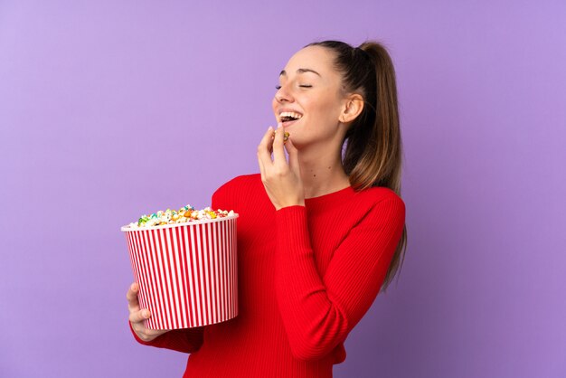 Giovane donna del brunette sopra la parete viola isolata che tiene un grande secchio di popcorn
