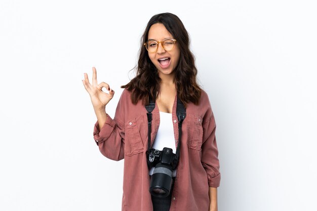 Giovane donna del fotografo sopra la parete bianca che mostra segno giusto con le dita