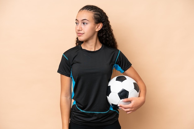 Giovane donna del giocatore di gioco del calcio isolata su fondo beige che guarda al lato