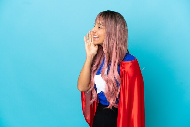 Giovane donna del supereroe con capelli rosa isolata su fondo blu che grida con la bocca spalancata al lato