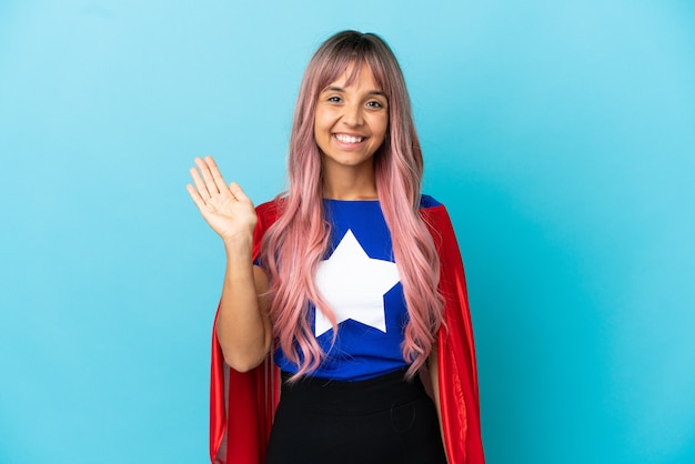 Giovane donna del supereroe con capelli rosa isolata su fondo blu che saluta con la mano con l'espressione felice