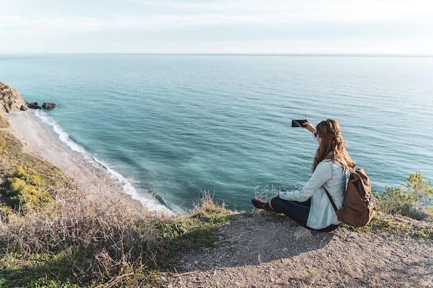 Giovane donna dell'anca con uno zaino che esplora e che fotografa la costa un bello giorno. Concetto di esplorazione e avventure