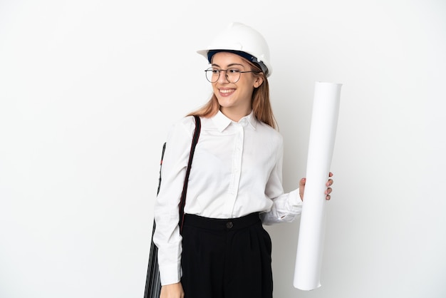 Giovane donna dell'architetto con il casco e che tiene i modelli isolati sulla parete bianca che osserva al lato e che sorride