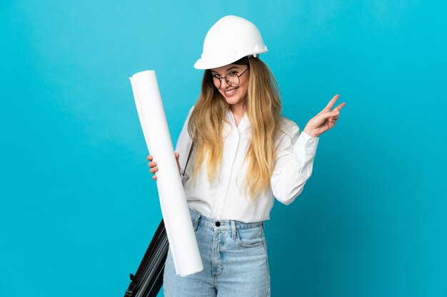 Giovane donna dell'architetto con il casco e che tiene i modelli isolati sulla parete blu che sorride e che mostra il segno di vittoria