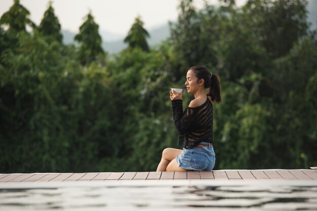 giovane donna dell'Asia che si siede nel parco su una degli alberi con una tazza di caffè.