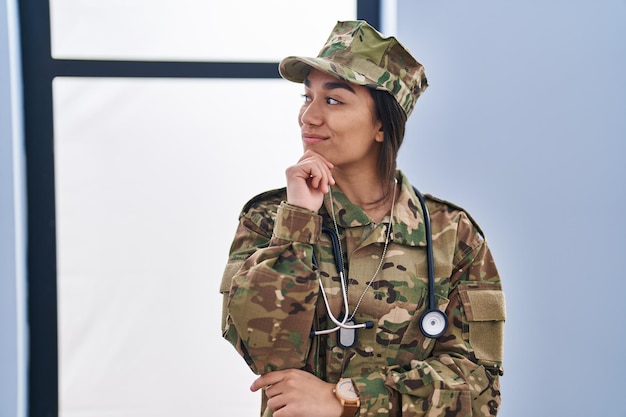 Giovane donna dell'Asia meridionale che indossa l'uniforme dell'esercito mimetico e il pensiero dello stetoscopio concentrato sul dubbio con il dito sul mento e alzando lo sguardo chiedendosi