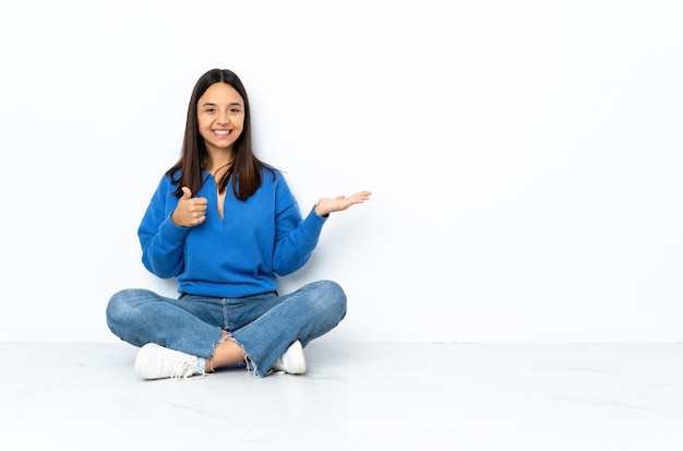 Giovane donna della corsa mista che si siede sul pavimento isolato sullo spazio bianco che tiene copyspace immaginario sulla palma per inserire un annuncio e con i pollici su