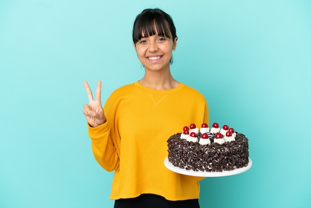 Giovane donna della corsa mista che tiene la torta di compleanno che sorride e che mostra il segno di vittoria