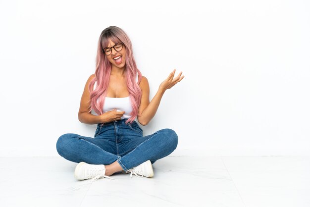 Giovane donna della corsa mista con capelli rosa che si siede sul pavimento isolato su fondo bianco che fa il gesto della chitarra