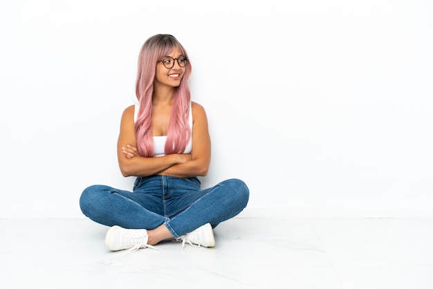 Giovane donna della corsa mista con capelli rosa che si siede sul pavimento isolato su fondo bianco che guarda side