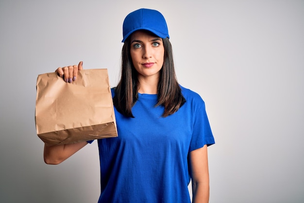 Giovane donna delle consegne con gli occhi azzurri che indossa il berretto che tiene il sacchetto di carta con il cibo con un'espressione sicura sul viso intelligente che pensa seriamente