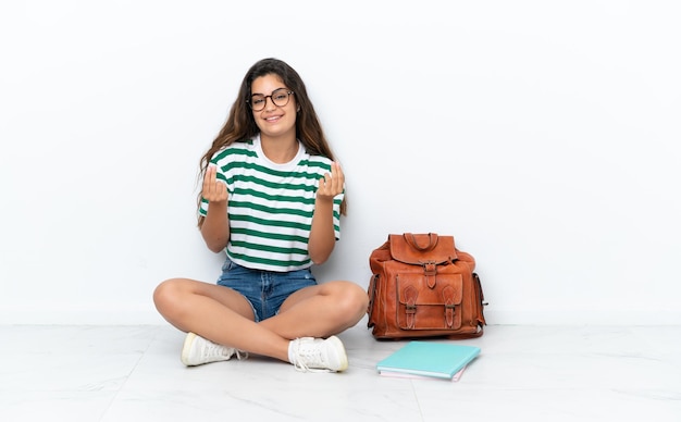 Giovane donna dello studente che si siede uno sul pavimento isolato su fondo bianco che fa il gesto dei soldi