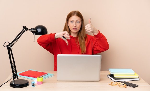 Giovane donna dello studente in un posto di lavoro con un computer portatile che fa segno buono-cattivo. Indeciso tra sì o no