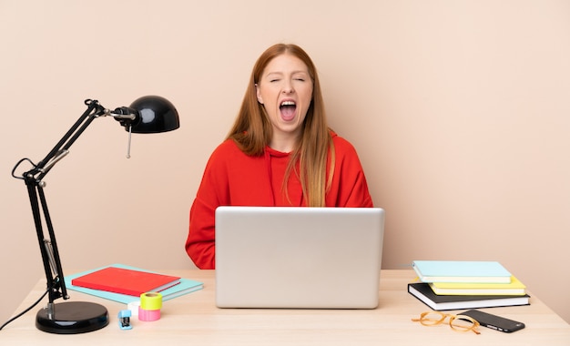 Giovane donna dello studente in un posto di lavoro con un computer portatile che grida alla parte anteriore con la bocca spalancata