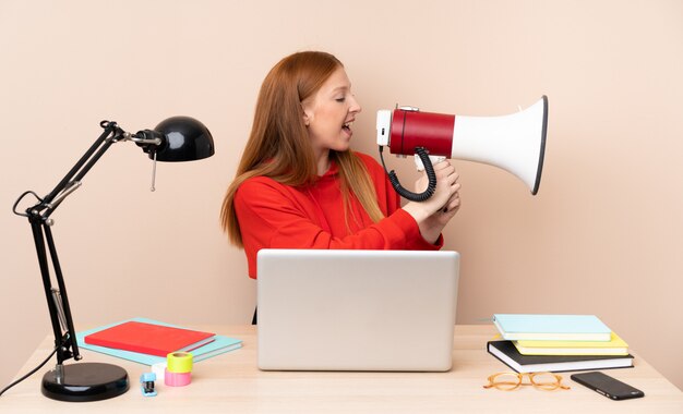 Giovane donna dello studente in un posto di lavoro con un computer portatile che grida tramite un megafono