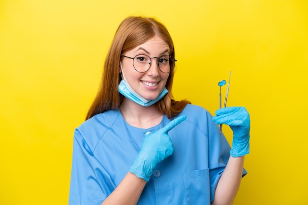 Giovane donna dentista rossa isolata su sfondo giallo che indica il lato per presentare un prodotto