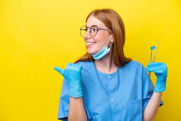 Giovane donna dentista rossa isolata su sfondo giallo che indica il lato per presentare un prodotto