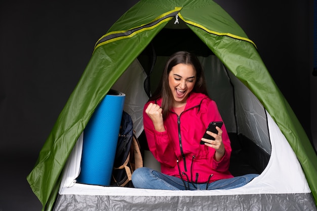 Giovane donna dentro una tenda verde di campeggio con il telefono nella posizione di vittoria