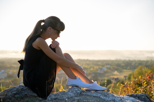 Giovane donna depressa in abito nero corto estivo seduto su una roccia pensando all'aperto al tramonto Donna alla moda che contempla in una calda serata nella natura