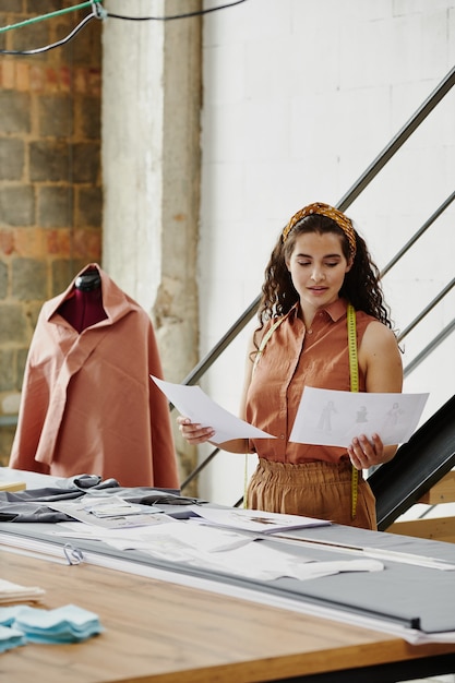 Giovane donna designer di abiti che guarda attraverso schizzi di nuovi modelli per la collezione stagionale e ne sceglie alcuni per il cucito