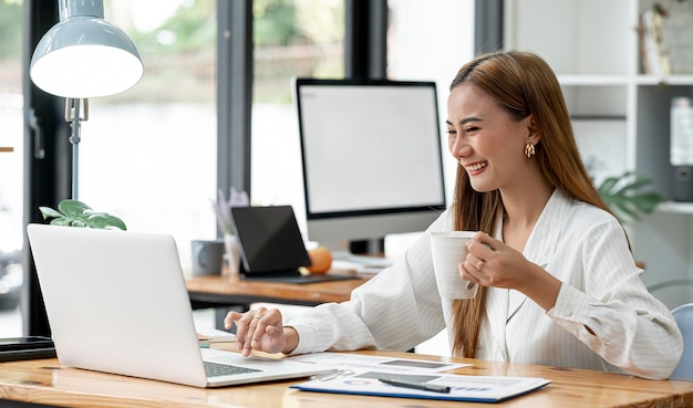 Giovane donna di affari asiatica bella tazza affascinante della holding, sorridente e lavorando al computer portatile in ufficio. Seduto a tavola.