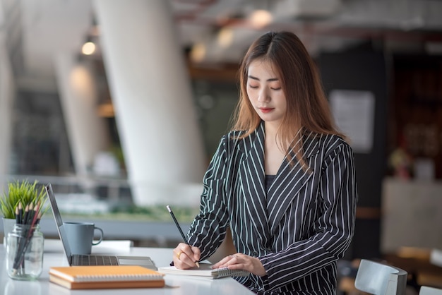 Giovane donna di affari asiatica che lavora al computer portatile e che prende nota mentre sedendosi al tavolo in ufficio