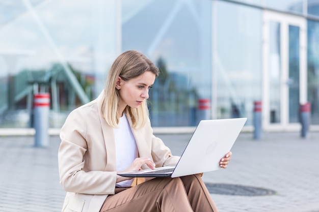 Giovane donna di affari che si siede sul marciapiede e che lavora al fondo urbano del computer portatile del centro moderno dell'ufficio. Donna d'affari, studentessa universitaria o universitaria online lavora all'aperto. Per le strade della città