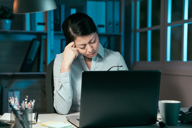 giovane donna di affari cinese asiatica stanca che lavora al computer portatile fino a notte. Ritratto di signora stressata casual con mal di testa che tiene occhiali e massaggio alla scrivania. Stile di vita dipendente femminile esausto