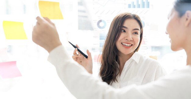 Giovane donna di affari creativa che scrive sulle note appiccicose su una parete di vetro che guarda collega femminile