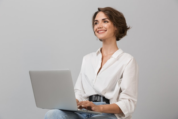 Giovane donna di affari in posa isolato sopra il muro grigio seduto su uno sgabello utilizzando il computer portatile