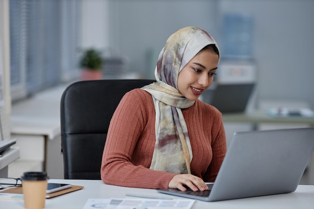 Giovane donna di affari musulmana sorridente in velo facendo uso del computer portatile