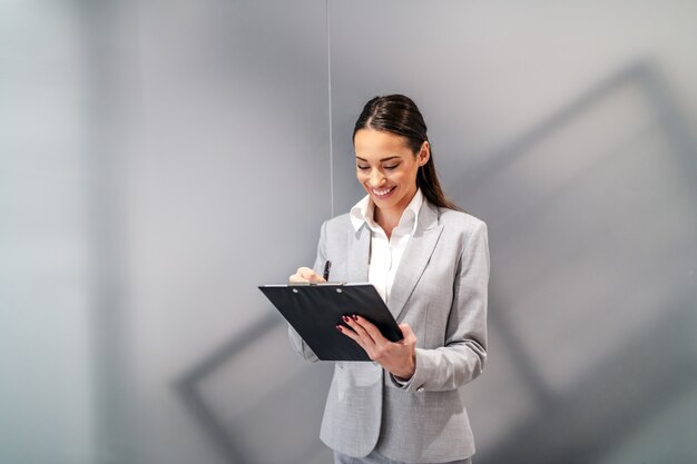 Giovane donna di affari sorridente caucasica attraente che si leva in piedi all'interno dell'azienda corporativa e che riempie i documenti sui appunti.