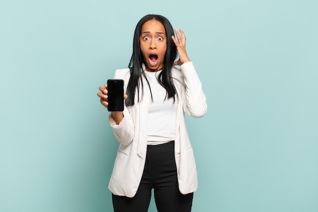 Giovane donna di colore che grida con le mani in alto in aria, sentendosi furiosa, frustrata, stressata e sconvolta. concetto di smart phone
