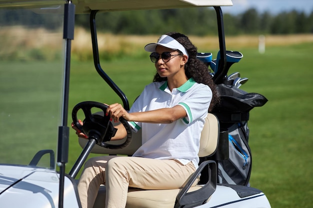 Giovane donna di colore che guida il carrello da golf sul campo verde alla luce del sole