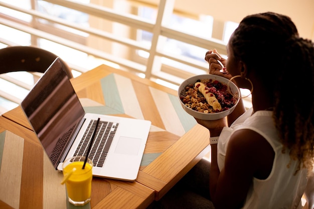 Giovane donna di colore che mangia una sana colazione mentre lavora al computer portatile nella caffetteria