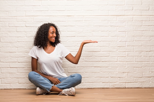 Giovane donna di colore che si siede su un pavimento di legno che tiene qualcosa con le mani, mostrando un prodotto, sorridente e allegro, offrendo un oggetto immaginario