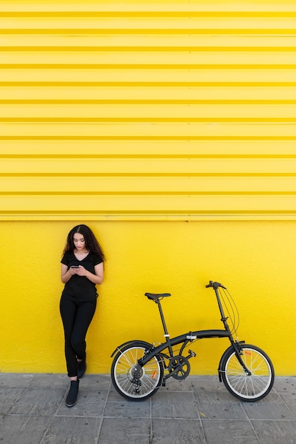 Giovane donna di colore con i capelli lunghi e vestito nero che manda un sms a qualcuno accanto alla sua bicicletta davanti a un muro giallo