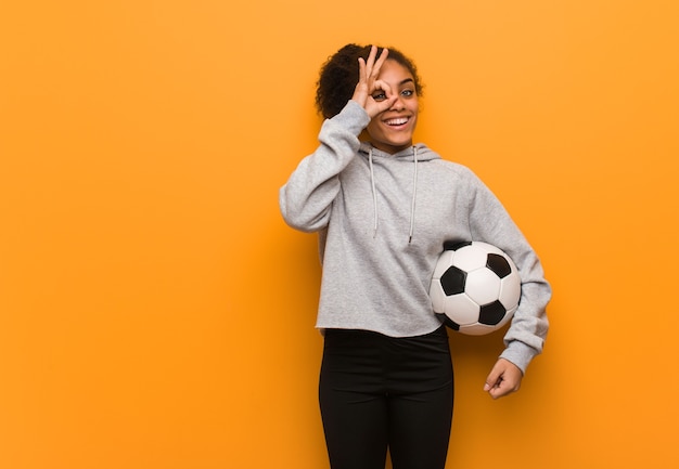 Giovane donna di colore fitness fiducioso facendo gesto ok sull'occhio. Tenendo un pallone da calcio.