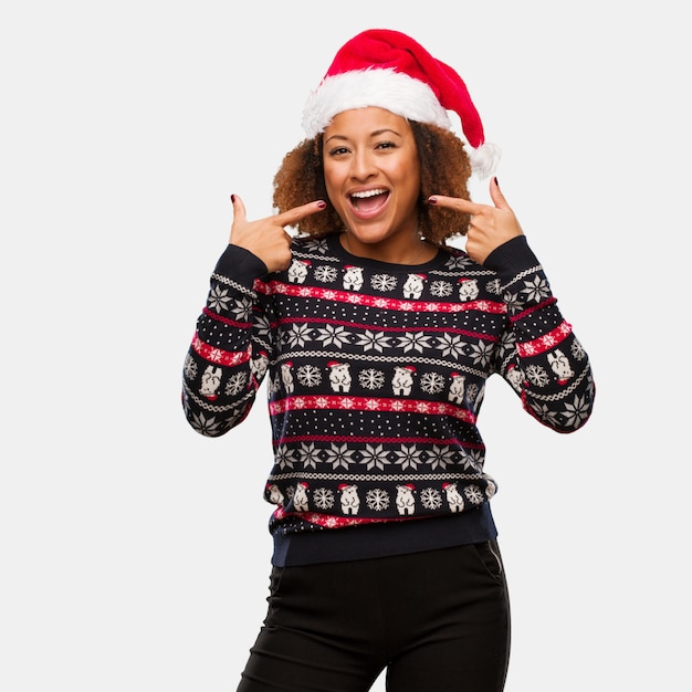 Giovane donna di colore in un maglione di Natale alla moda con sorrisi di stampa, che indica la bocca