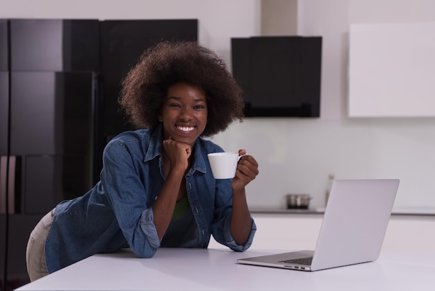 Giovane donna di colore sorridente che per mezzo del computer e bevendo caffè nell&#39;interno moderno della cucina