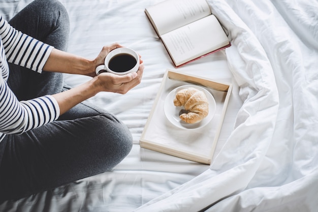 Giovane donna di felicità sul letto con il vecchio libro e la tazza di caffè di mattina in mani e Croi