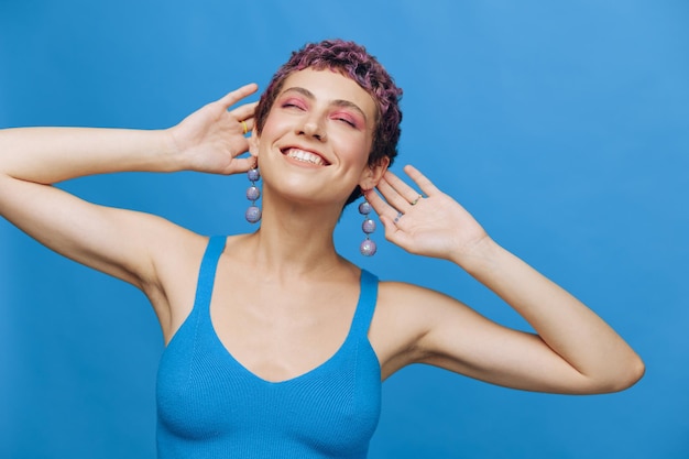 Giovane donna di moda atletica con capelli colorati e taglio di capelli corto che posa e balla in abbigliamento sportivo blu sorridendo e guardando il primo piano della fotocamera su uno sfondo blu monocromatico