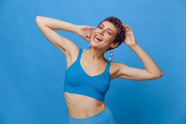 Giovane donna di moda atletica con capelli colorati e taglio di capelli corto che posa e balla in abbigliamento sportivo blu sorridendo e guardando la telecamera su uno sfondo blu monocromatico