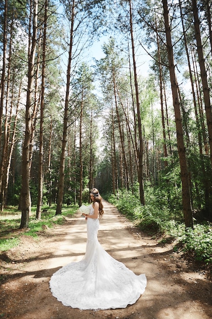 Giovane donna di modello in abito da sposa in pizzo elegante mantenendo un bouquet e in posa nella foresta.