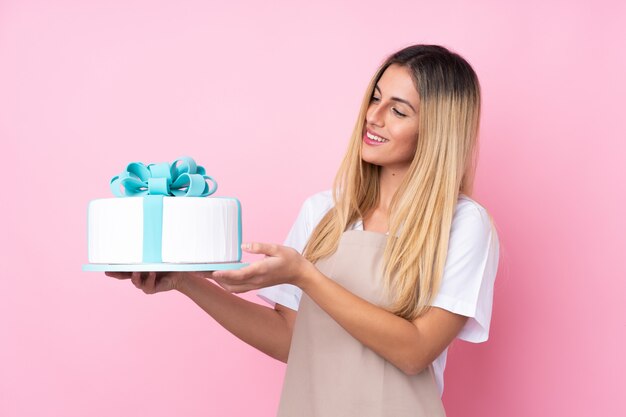 Giovane donna di pasticceria uruguaiana con una grande torta sul muro rosa isolato con espressione felice