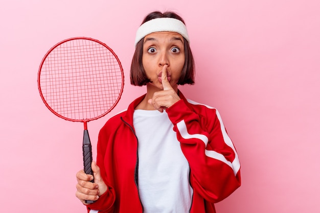 Giovane donna di razza mista che gioca a badminton isolato sul muro rosa mantenendo un segreto o chiedendo silenzio.