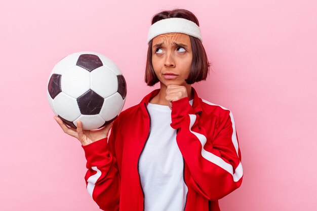 Giovane donna di razza mista che gioca a calcio isolato sul muro rosa che guarda lateralmente con espressione dubbiosa e scettica.